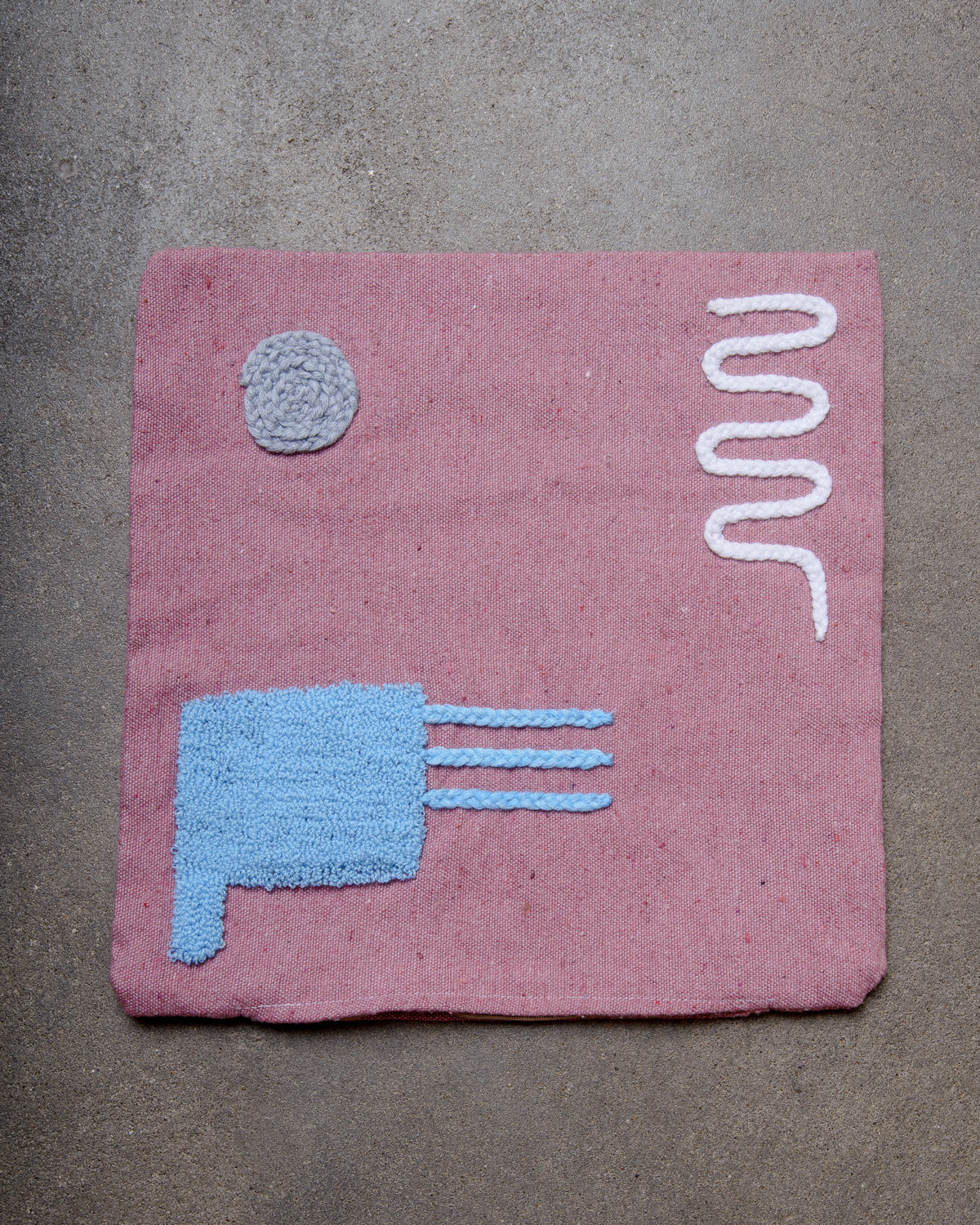 Pink wool cushion cover with embroidered design laying on concrete floor.